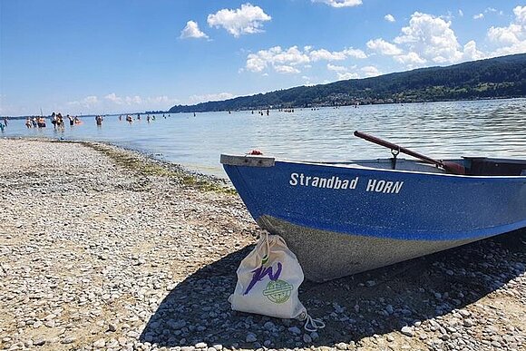 Ein blaues Holzboot vor dem Bodensee mit der Aufschrift Strandbad Horn davor ein Jutebeutel mit Logo