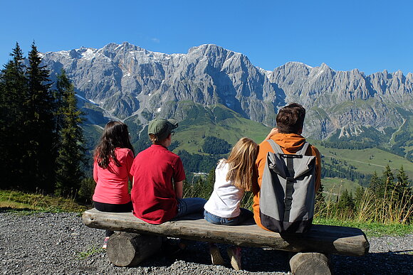 junge Menschen sitzen auf einem Baumstamm mit Bergpanorama im Hintergrund bei blauem Himmel