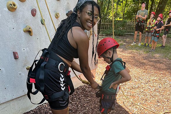 Person mit Kletterausrüstung an und Kind mit rotem Helm im Klettergarten